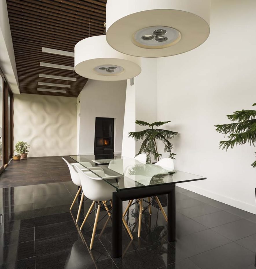 Modern glass dining table with white chairs in a spacious, elegantly designed room featuring wooden ceiling accents, plants, and a fireplace, reflecting Rizo Homes' commitment to quality residential design.
